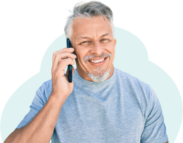 A man with silver hair swept across his forehead is smiling while talking on the phone. He wears a soft blue jersey cotton shirt and looks happy to be with the conversation.